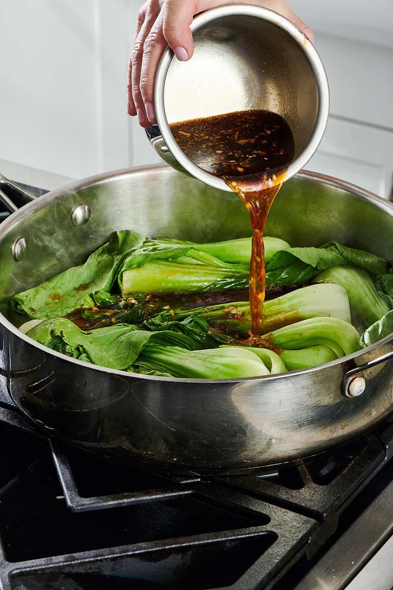 Soy sauce and broth mixture being poured over Baby Bok Choy in a pan.