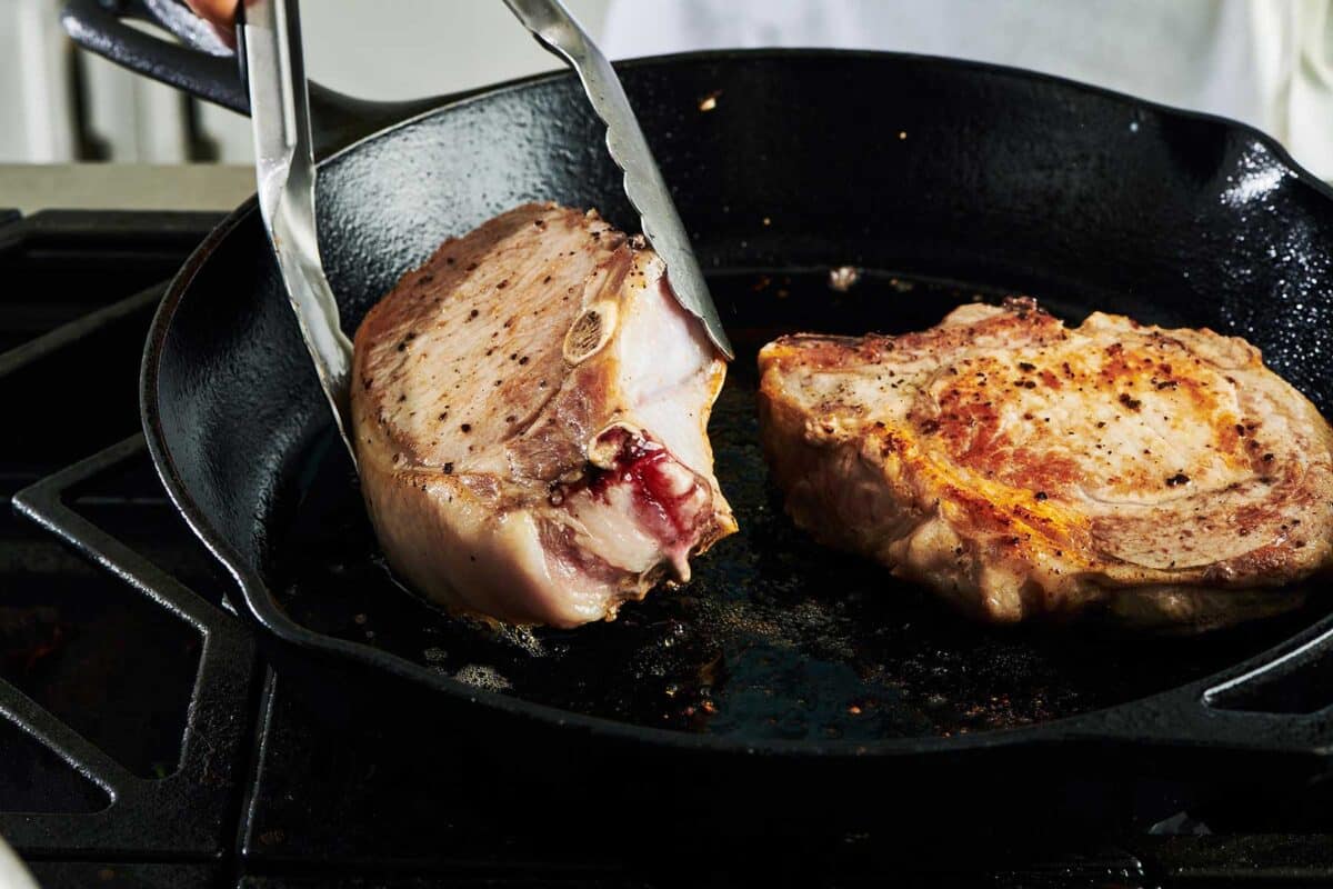 Flipping pork chops in cast iron pan on stove with tongs.