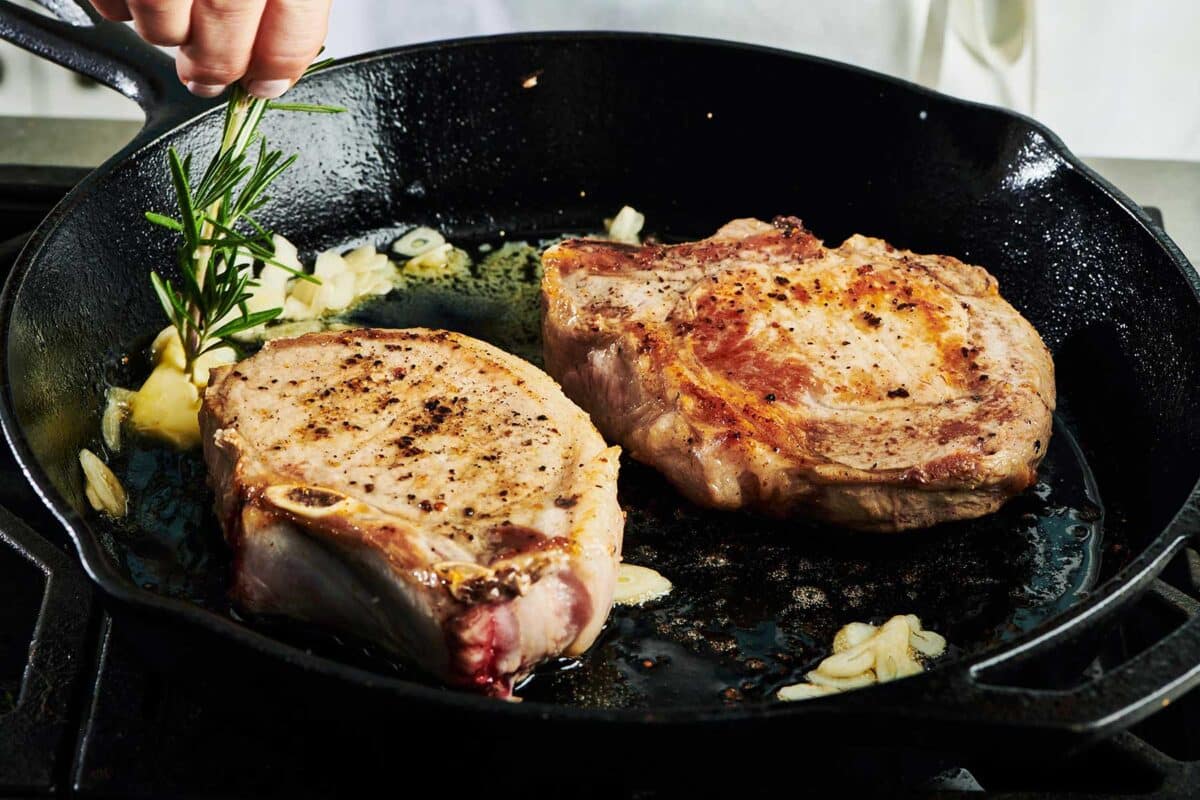 Adding fresh rosemary to pork chops in cast iron pan on stove.