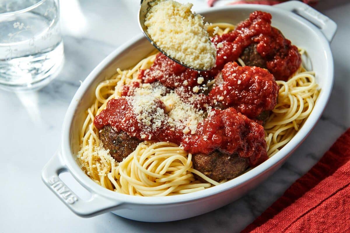 Spoon sprinkling Parmesan cheese onto pasta and meatballs.