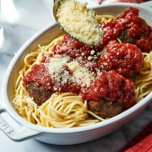 Spoon sprinkling Parmesan cheese onto pasta and meatballs.
