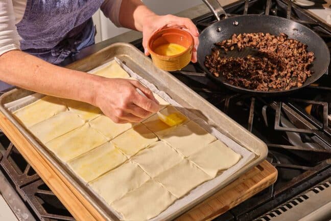 Woman brushing a cut sheet of puff pastry with beaten egg.