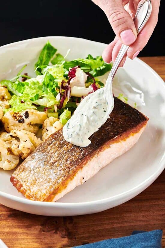 Spooning dill sauce on salmon fillet with crispy skin