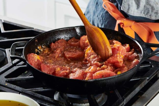 Stirring a pan of stewing tomatoes in a cast-iron pan on the stovetop.