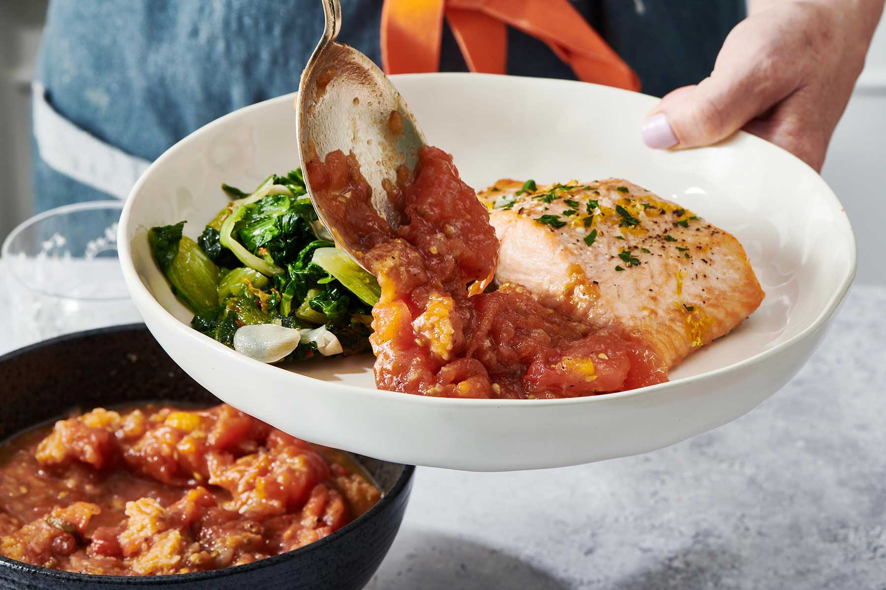 Woman serving stewed tomatoes on plate with fish filet and sautéed greens.