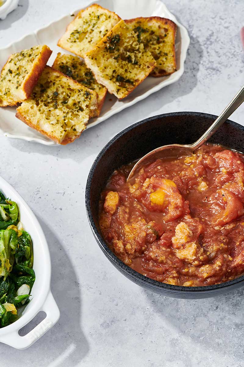 Stewed Tomatoes in black bowl in a table setting with garlic bread and cooked greens.