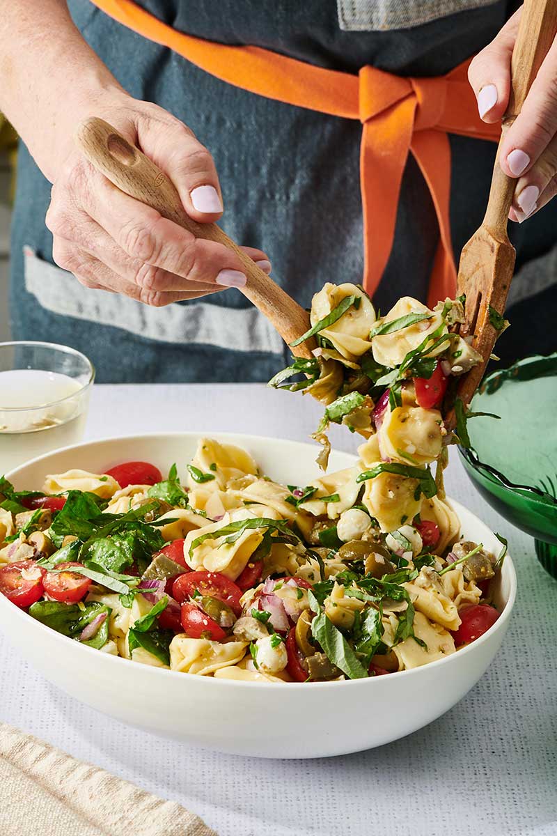 Woman tossing a bowl of Tortellini Pasta Salad with wood utensils.
