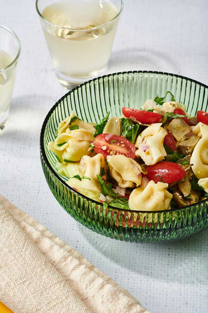 Tortellini Pasta Salad in a green glass bowl with glasses of wine on a table.