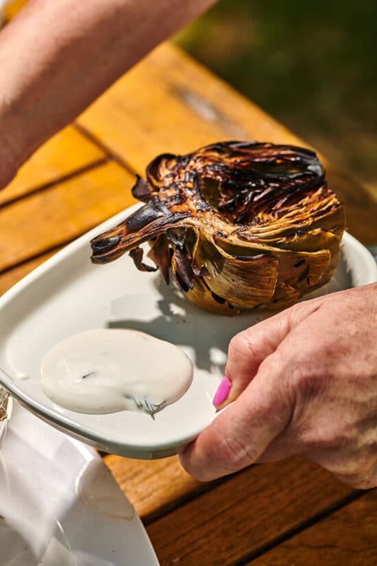 Woman holding plate of grilled artichoke and creamy lemon sauce at picnic table.