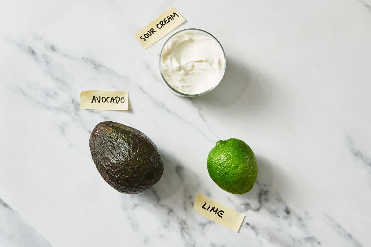 Avocado, lime, and sour cream to make Avocado Crema on white marble counter.