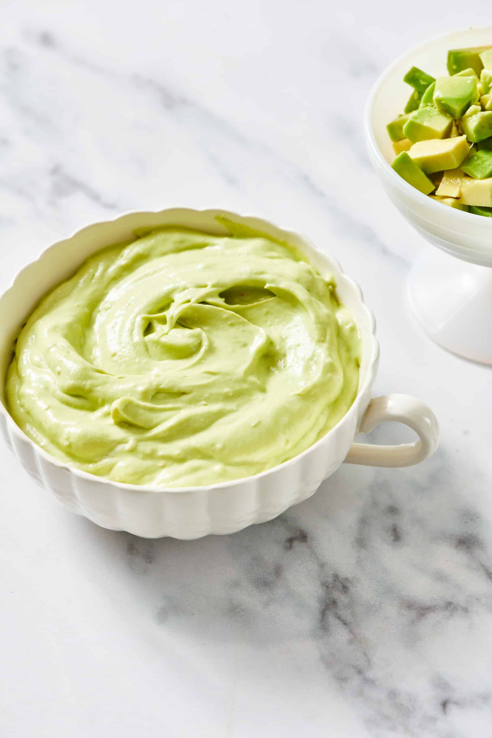 White crock of Avocado Crema on marble table with bowl of chopped avocado.