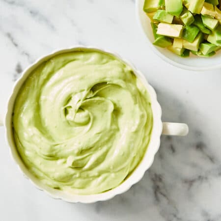 Fresh Avocado Crema in white crock with bowl of avocado on white marble counter.