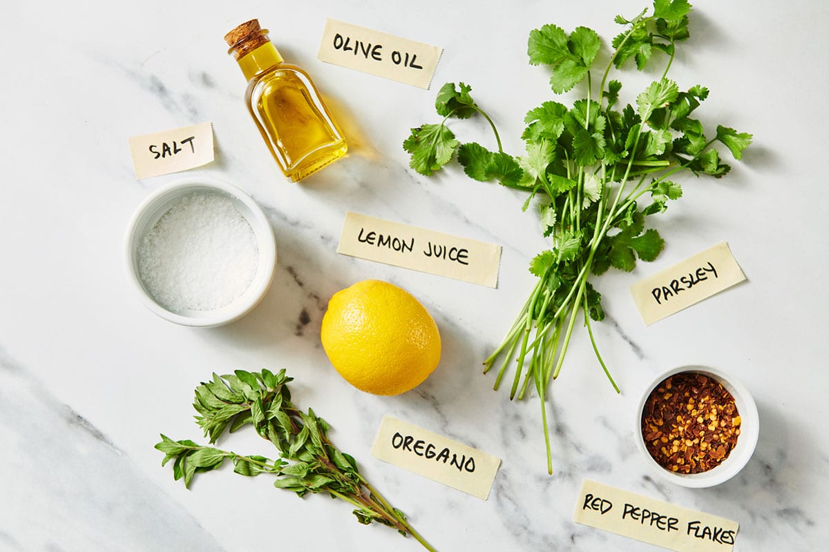 Ingredients like parsley, oregano, and lemon for Chimichurri Sauce on white marble counter