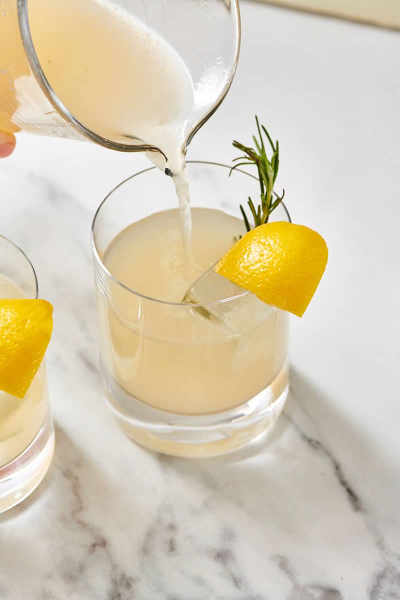 Pouring Lemongrass Cocktail over ice into glasses garnished with rosemary and lemon.