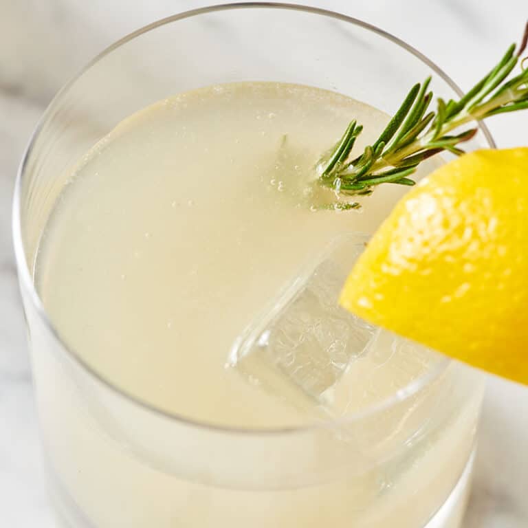 Lemongrass cocktail in glass with fresh rosemary and lemon wedge garnish on marble tabletop.