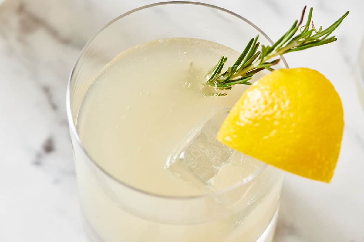 Lemongrass cocktail in glass with fresh rosemary and lemon wedge garnish on marble tabletop.