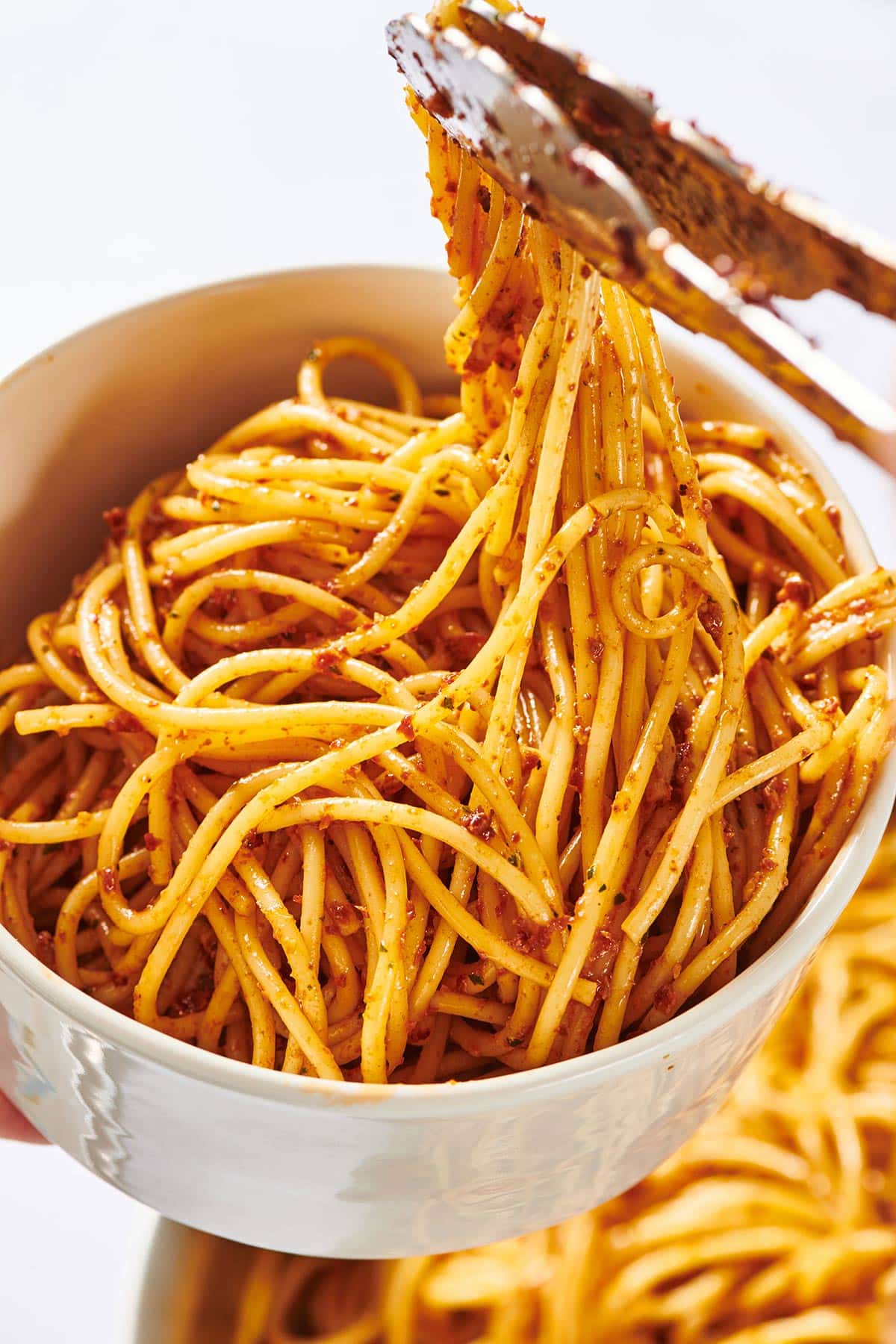 Tongs picking up spaghetti in sun-dried tomato pesto from bowl.