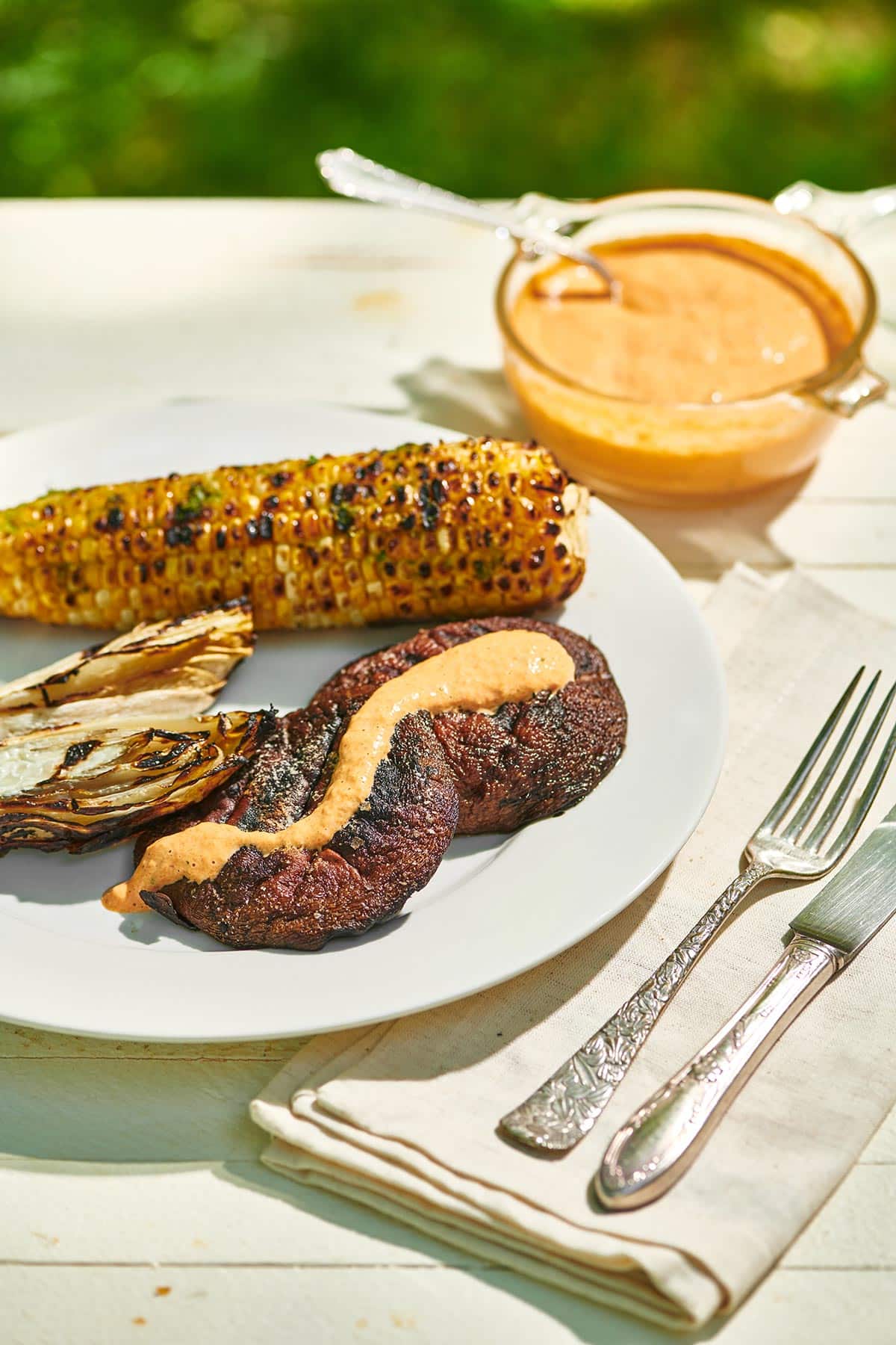 Grilled portobello mushrooms on white plate with grilled endive and corn and bowl of red pepper sauce.