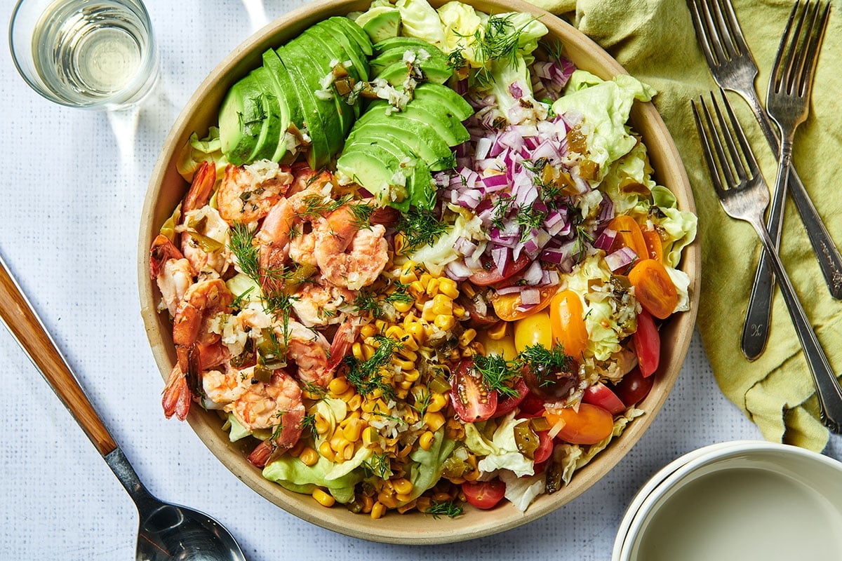 Brown bowl of Grilled Shrimp Salad with avocado on table with white wine.