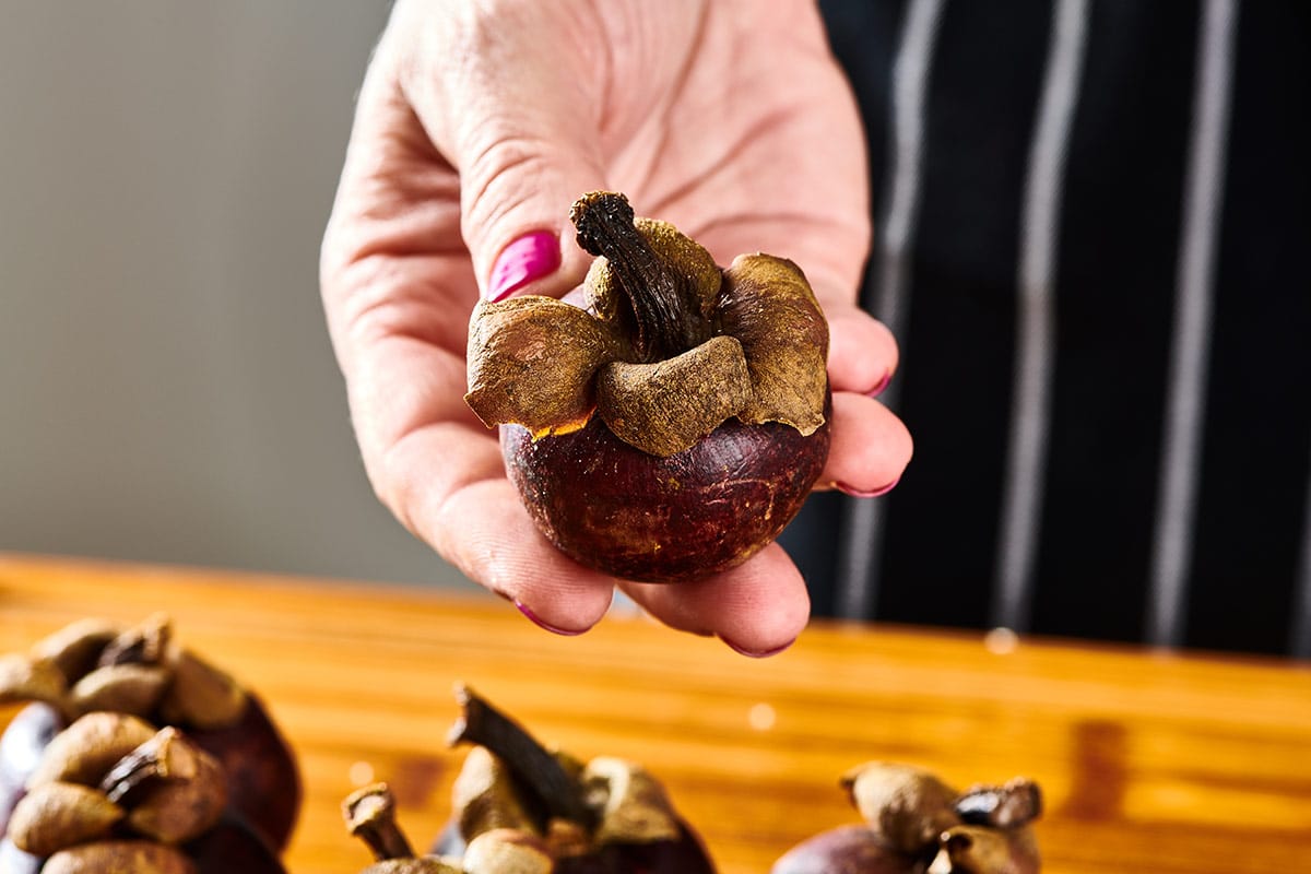 Woman holding whole mangosteen fruit in a kitchen over a wood cutting board.