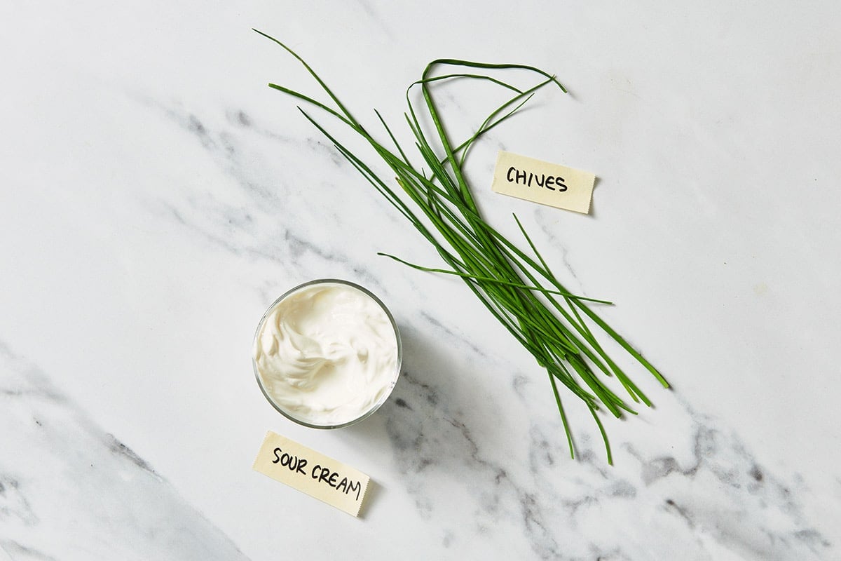 Sour cream in small dish next to fresh chives on white marble table.