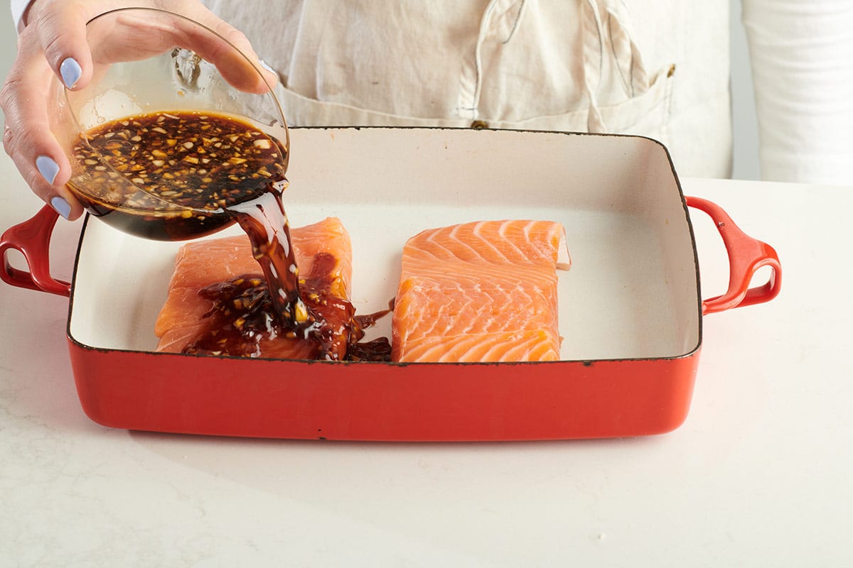 Woman pouring teriyaki sauce over salmon filets in red baking pan.