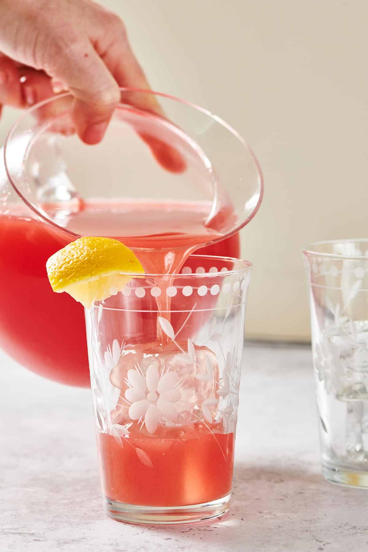 Pouring homemade watermelon lemonade into ice-filled glass with lemon wedge.