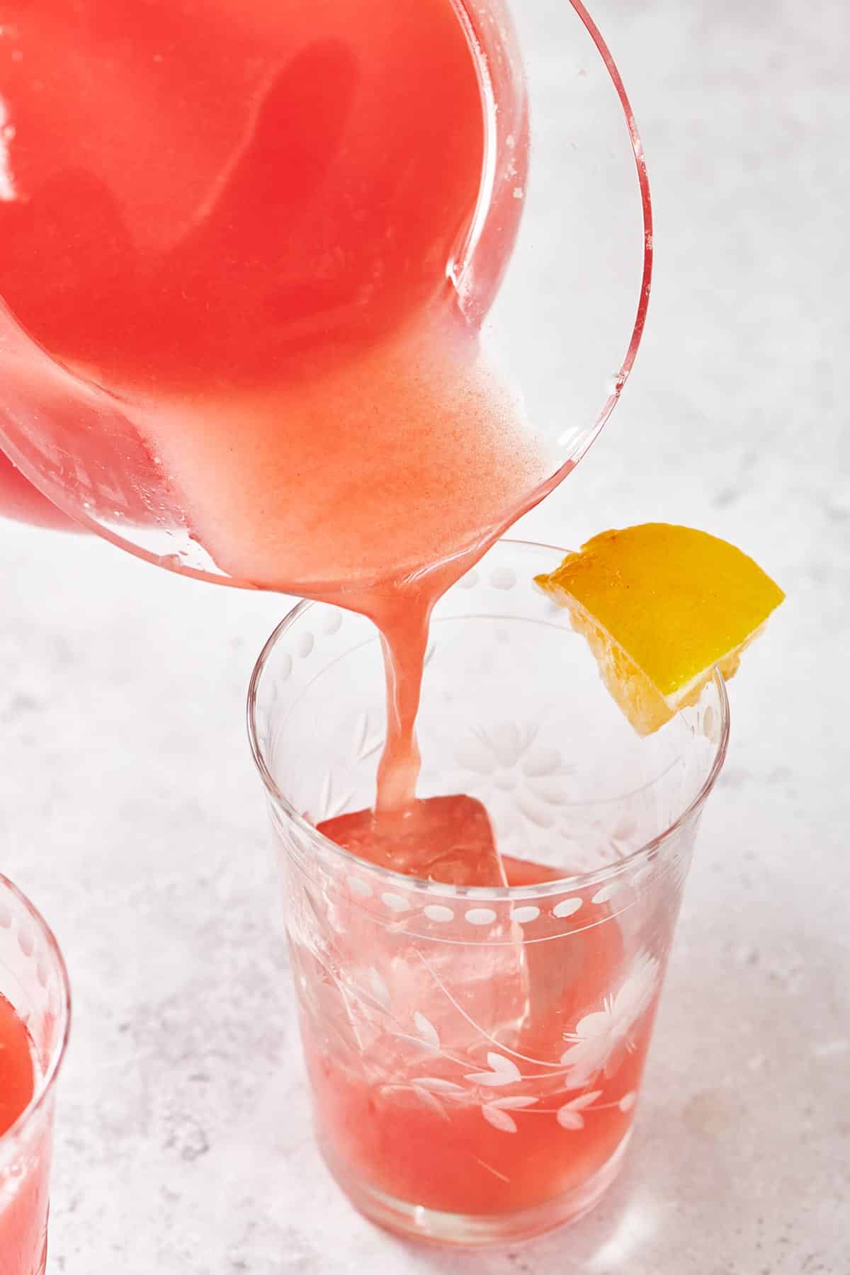 Pouring watermelon lemonade into ice-filled glass with lemon wedge.