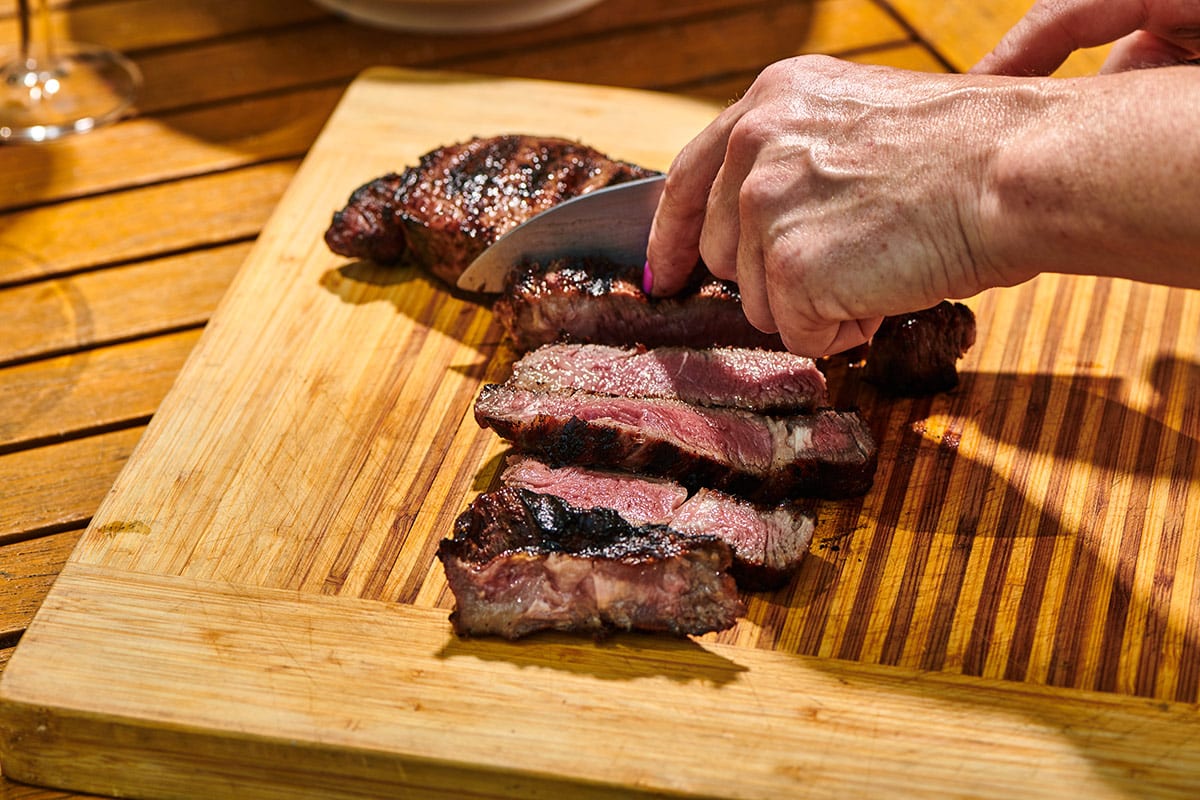 Slicing grilled rib-eye steak with knife on wood cutting board.