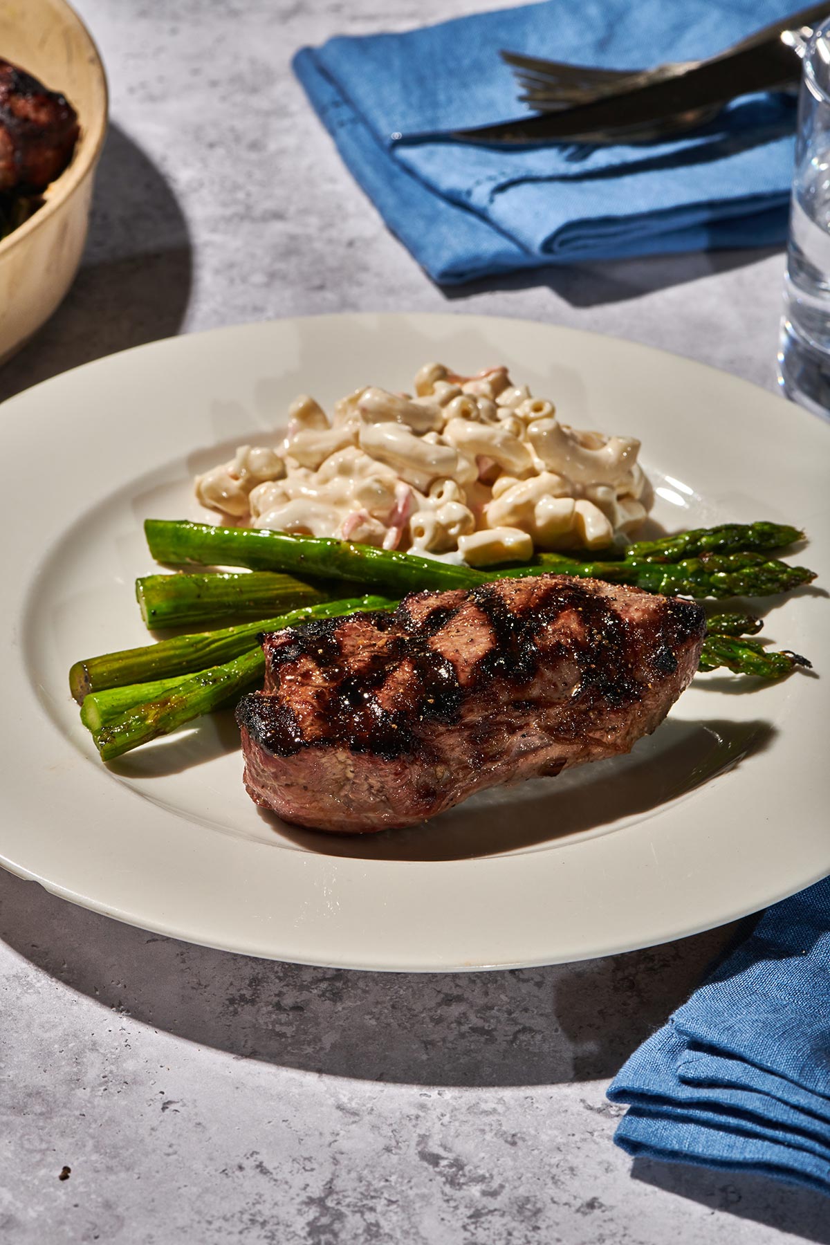 Cookout table with grilled top sirloin steak on plate with asparagus and macaroni salad.