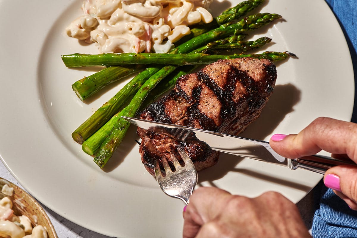 Slicing grilled top sirloin steak on plate with asparagus and macaroni salad.