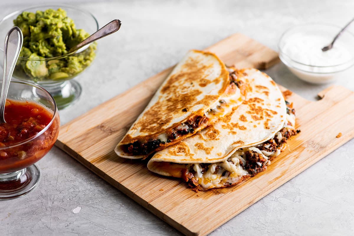 Beef quesadillas on wood cutting board near bowls of guacamole and salsa.
