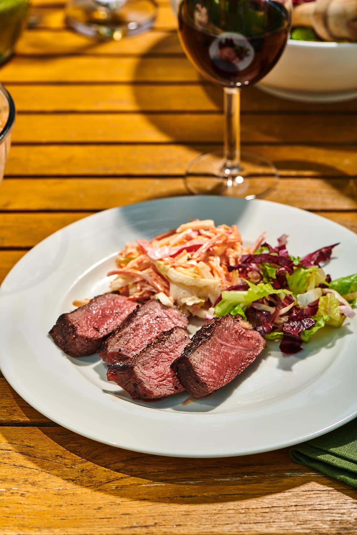 Outdoor table with plated ranch steaks and sides on white plate with glass red wine.