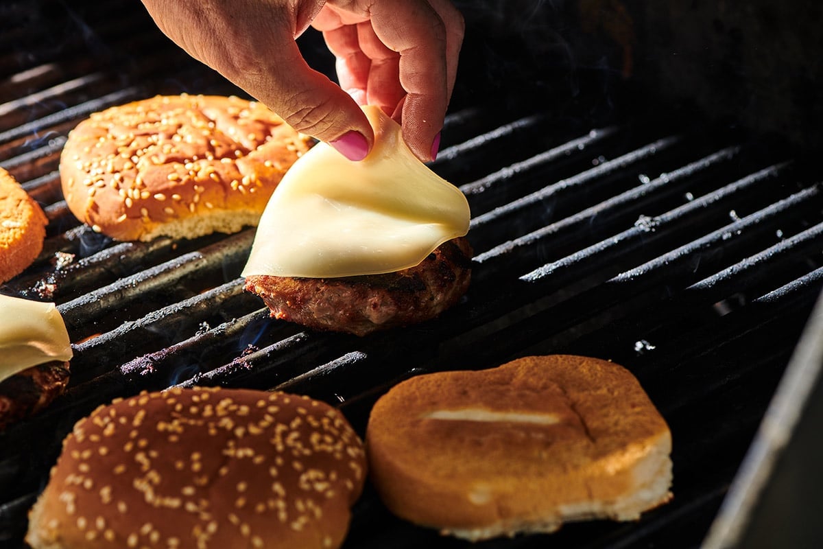 Woman placing cheese on turkey burger next to hamburger buns on grill.