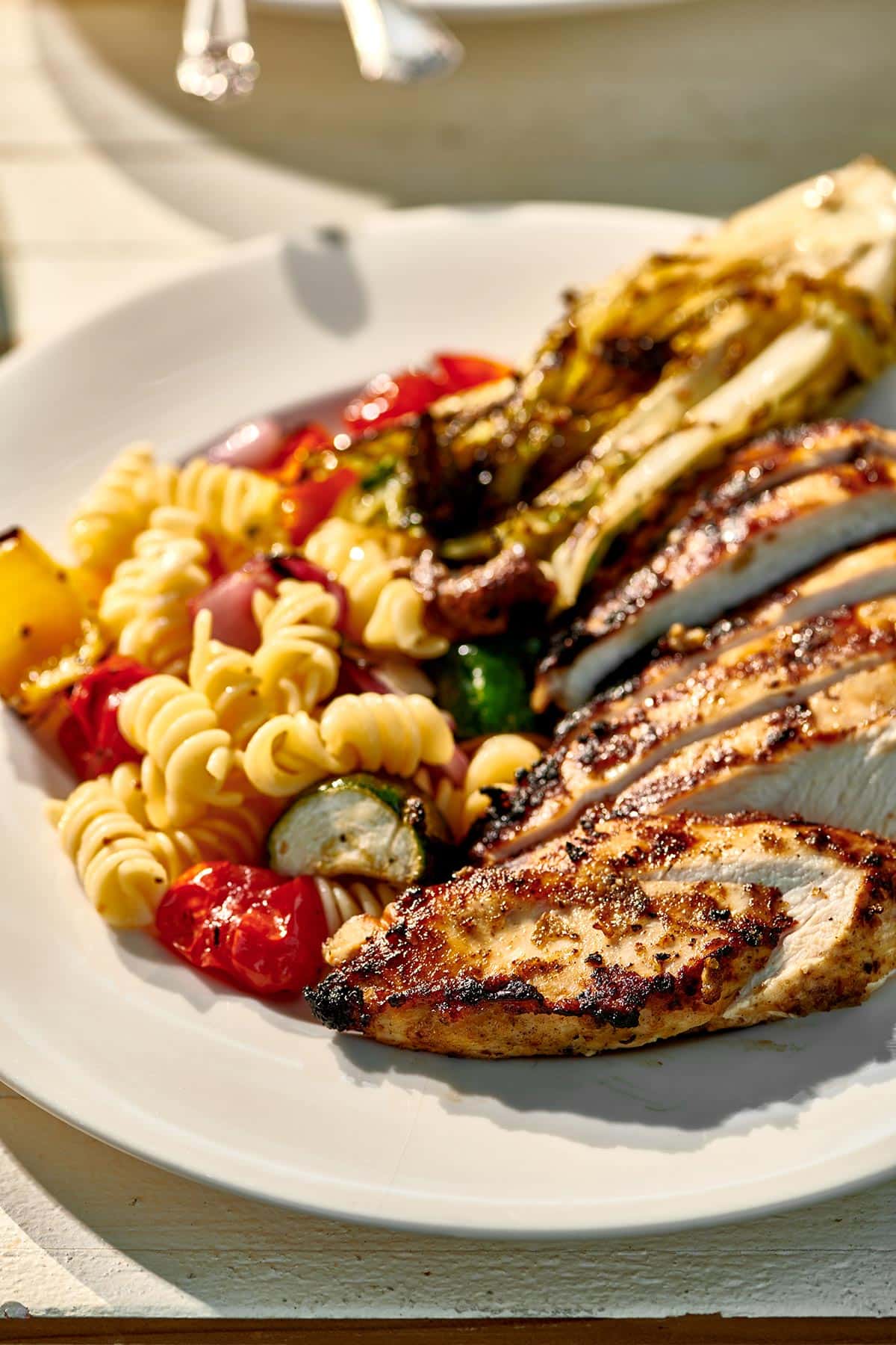 Plating slices of grilled chicken with veggie pasta salad at cookout.