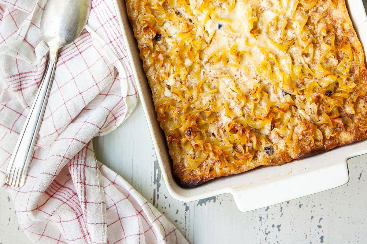 Noodle kugel in baking pan on table with towel and serving spoon.