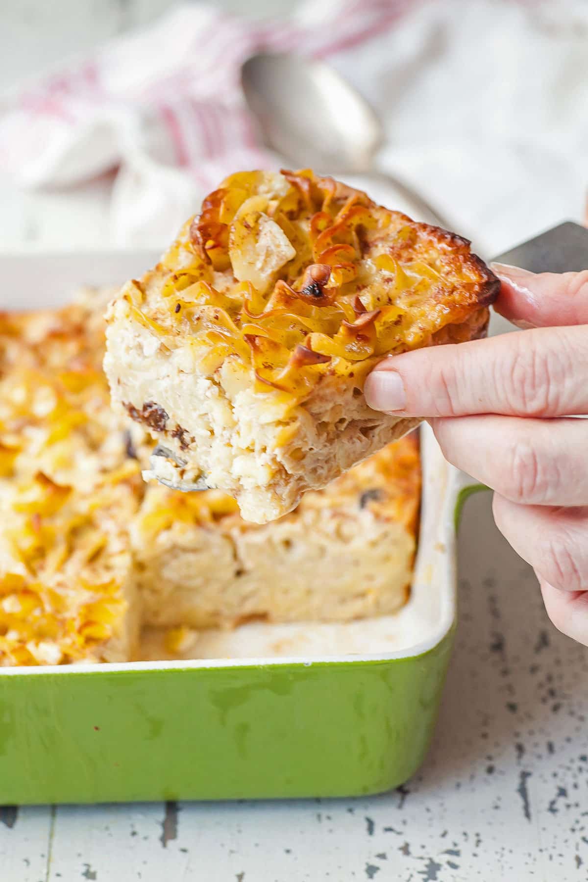 Pulling cut piece of freshly baked Jewish noodle kugel from green baking dish.