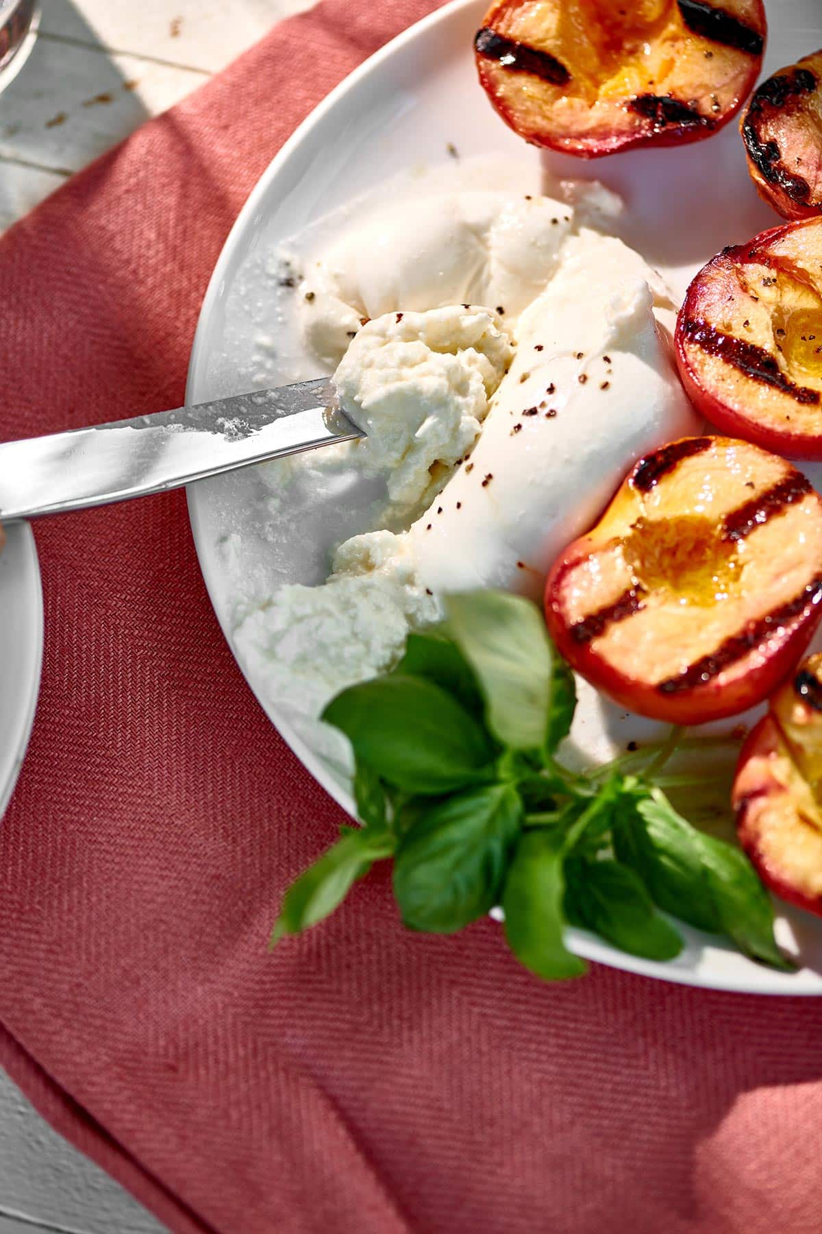 Cutting ball of burrata cheese with knife next to grilled peaches.