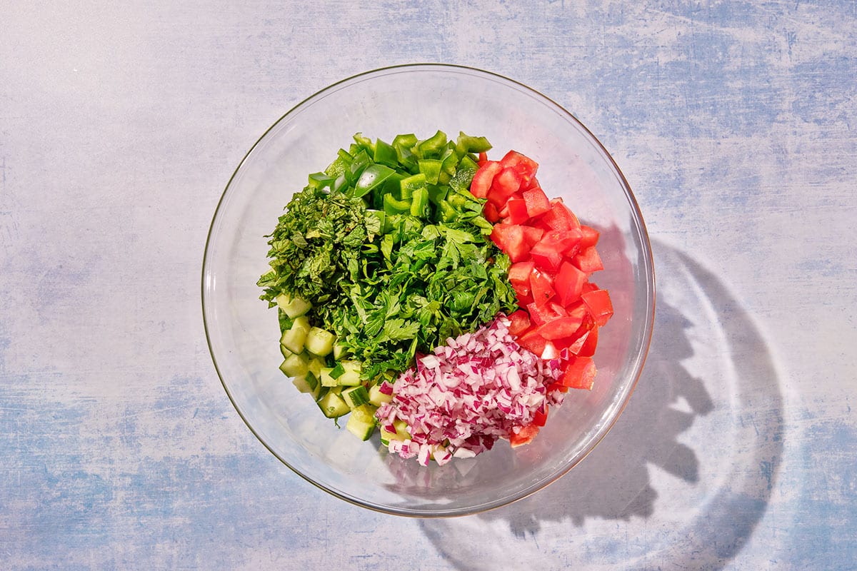 Chopped tomatoes, cucumbers, onions, and herbs in glass bowl.
