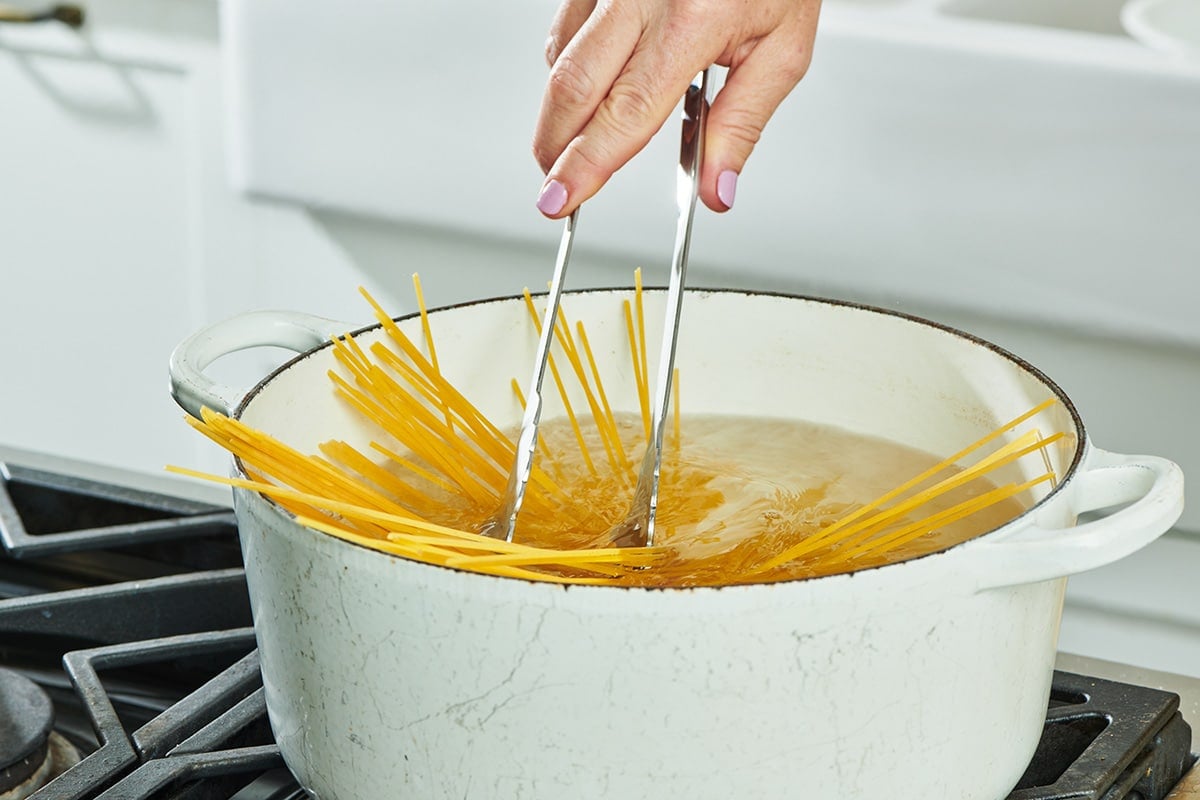 Stirring spaghetti in white pot of boiling water with tongs.