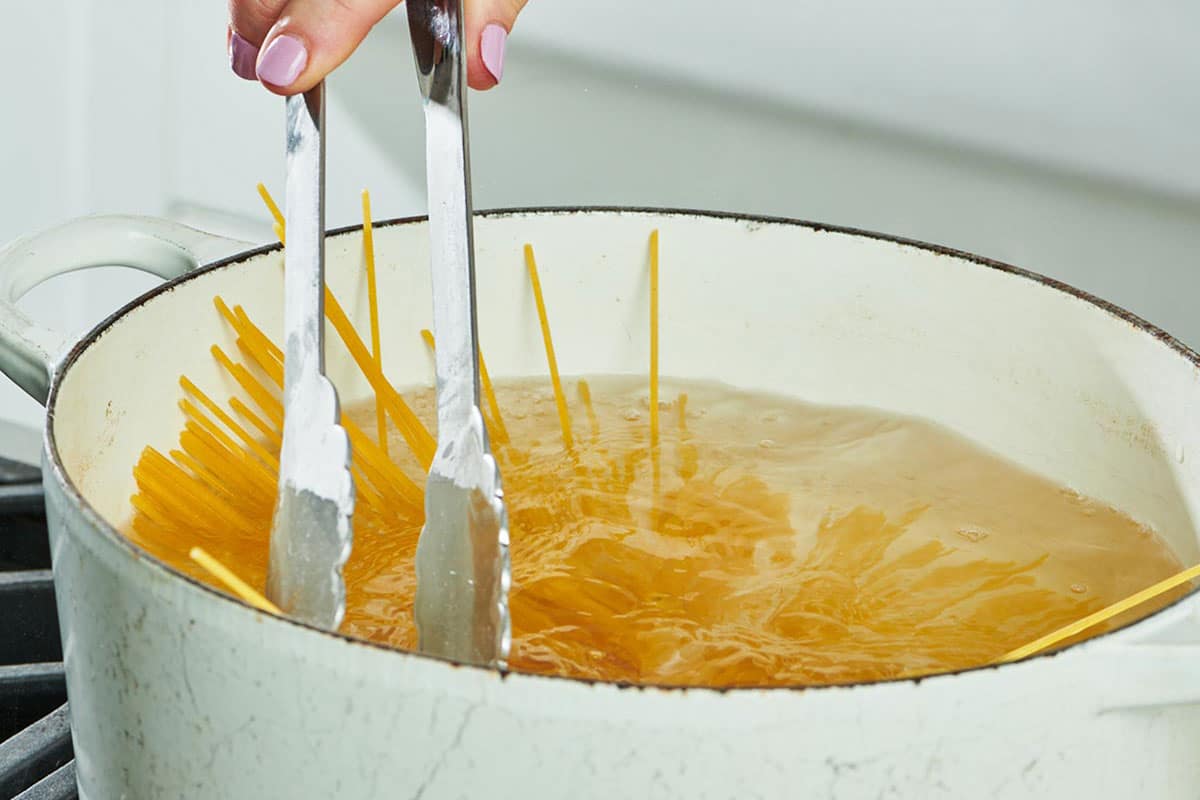 Stirring spaghetti in white pot of boiling water with tongs.