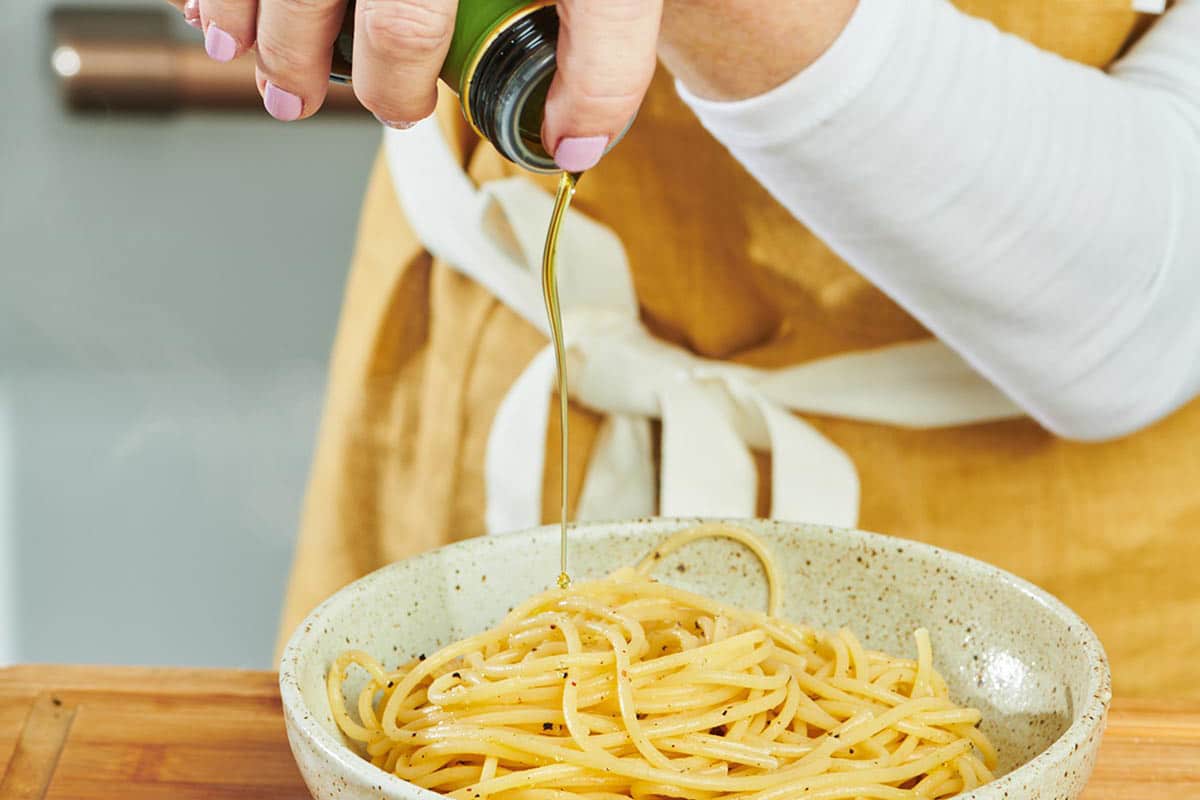 Woman adding oil to bowl of cooked spaghetti.
