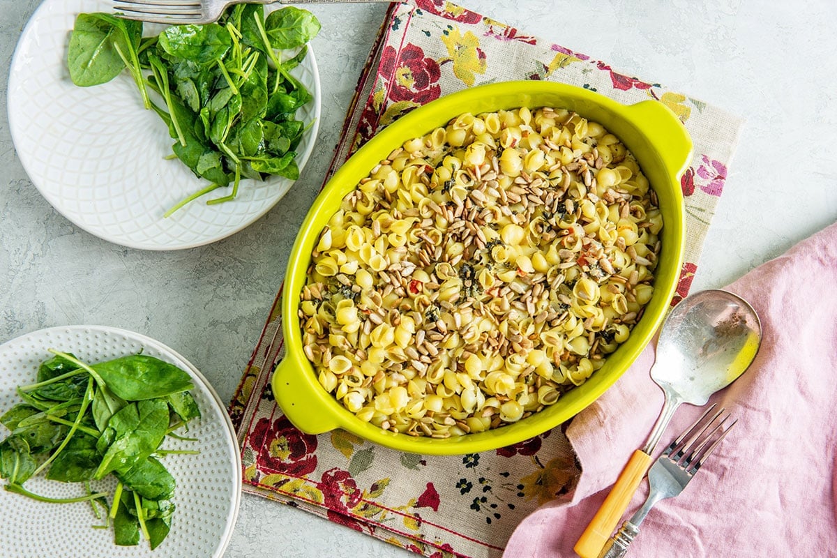 Spinach Goat Cheese Baked Pasta with Sunflower Seeds in yellow dish on table alongside plates with salads.