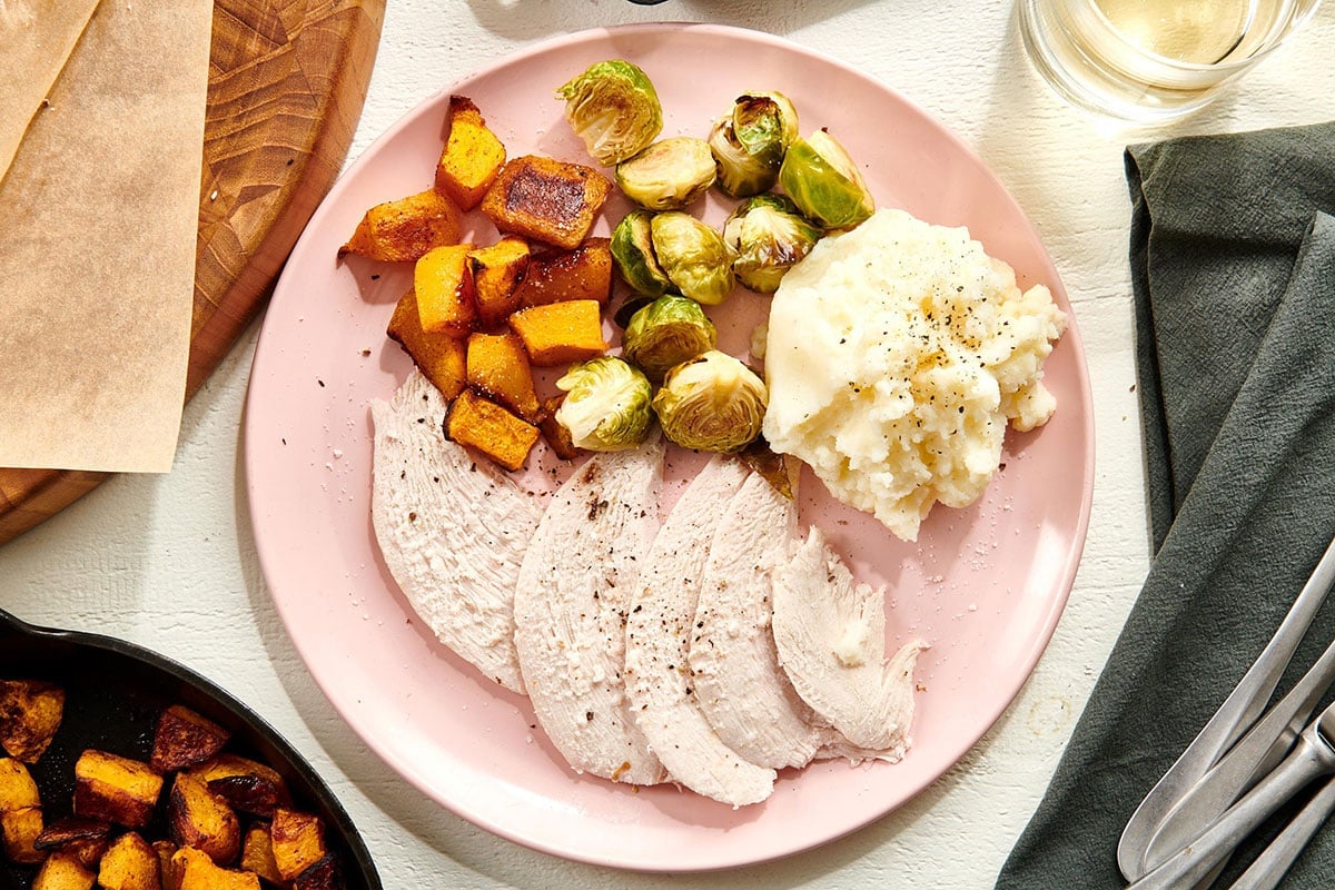 Sliced turkey breast on plate with sweet potatoes, Brussels sprouts, and mashed potatoes.