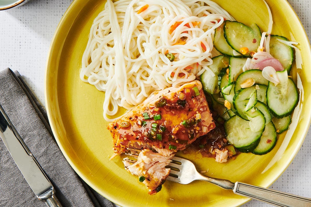 Fork on yellow plate with ginger lemongrass salmon, noodles, and cucumber salad.