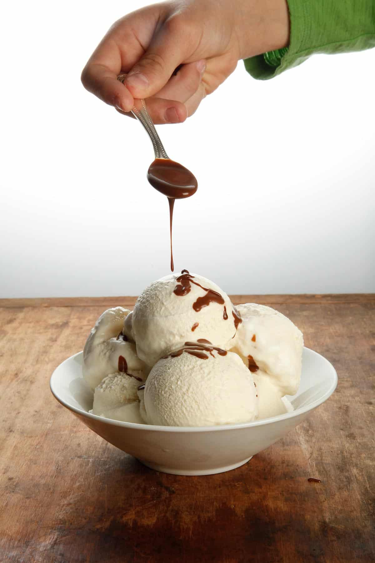 Pouring hot fudge sauce over bowl of vanilla ice cream on wood table.