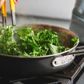 Stirring broccoli rabe with tongs as it cooks in large pan on stove.