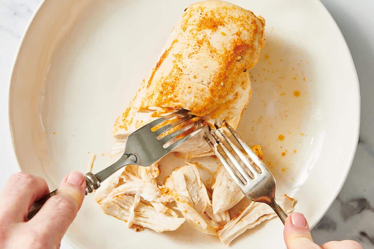 Shredding chicken breast cooked in instant pot with forks on white plate.