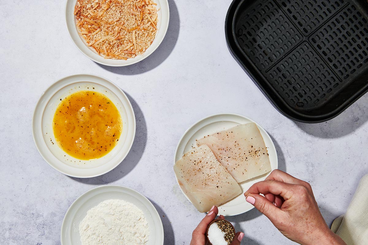 Seasoning halibut filets with salt and pepper on table with breading bowls and air fryer.