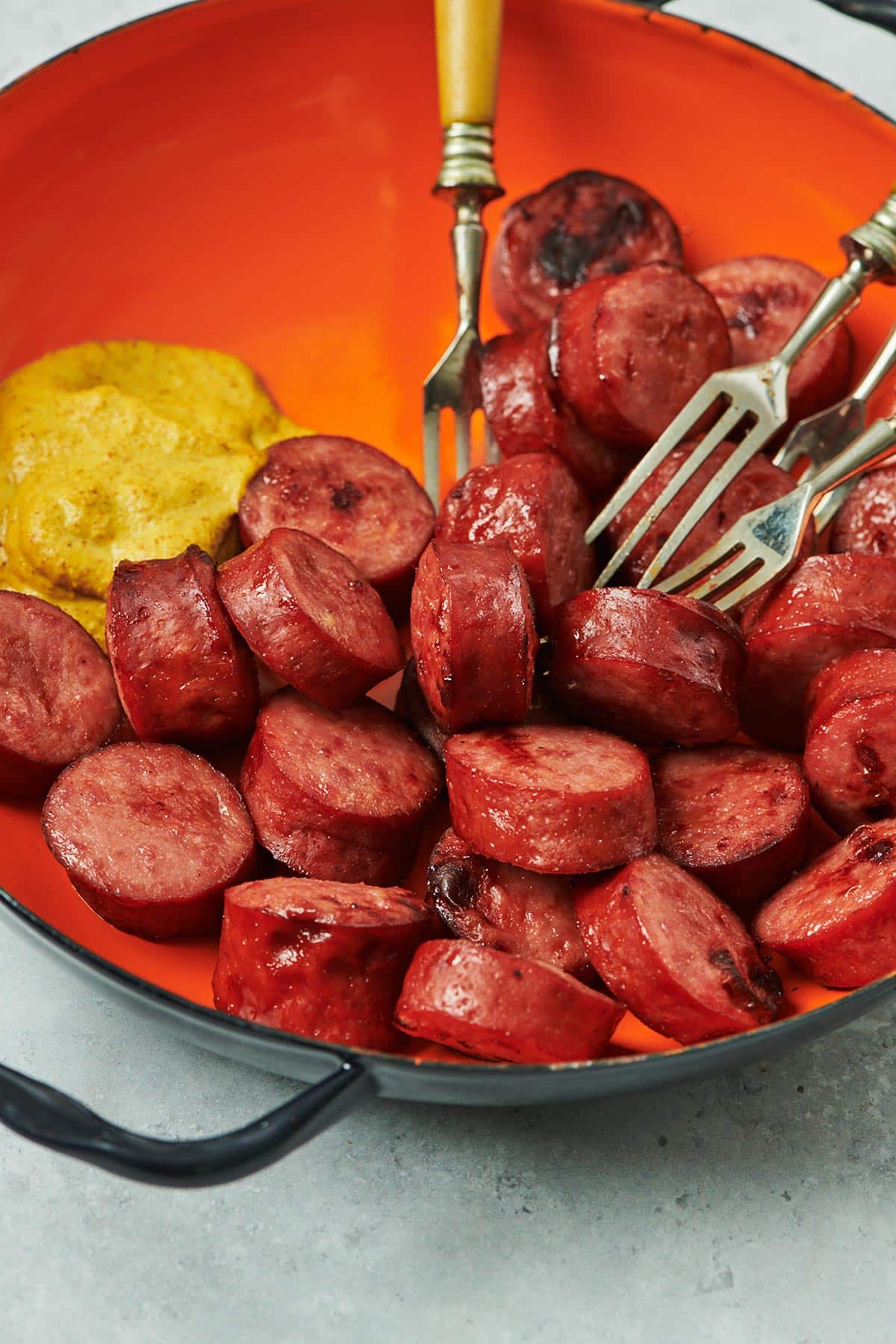 Sliced kielbasa in orange serving bowl with mustard and forks.